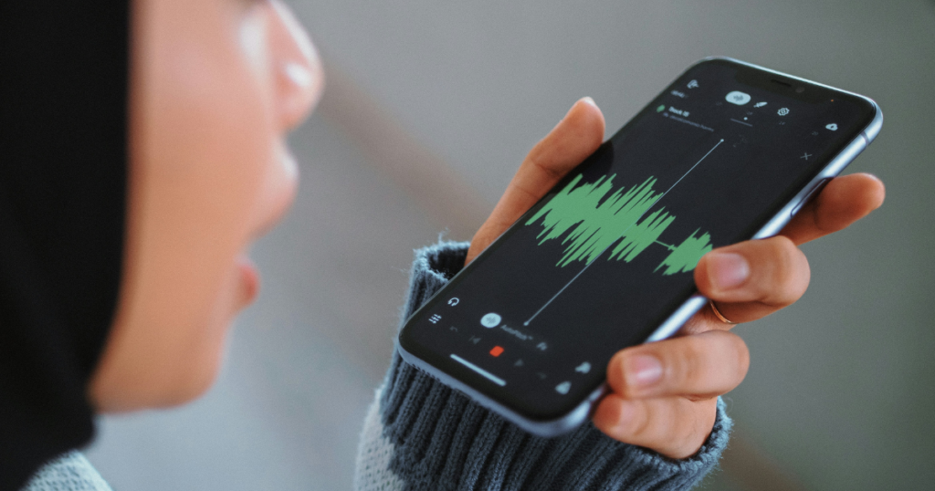 Close-up of a person in a hijab holding a smartphone displaying an active voice-waveform graphic, demonstrating the "voice-to-image" prompt feature found in a modern Artificial Intelligence photo generator.