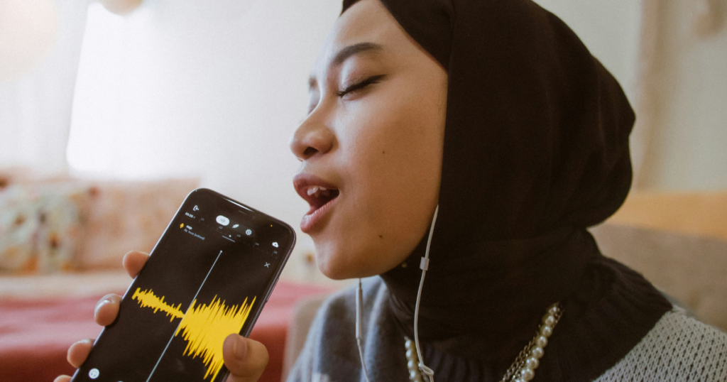 A young woman in a black hijab speaking into a smartphone with a visible yellow sound waveform on the screen,